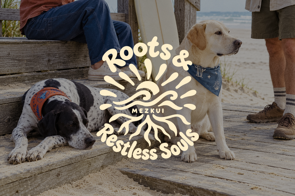 Dois cães com bandanas num passadiço à beira-mar com pessoas e uma prancha de surf, sob um emblema de cor creme Roots & Restless Souls MEZKUI.